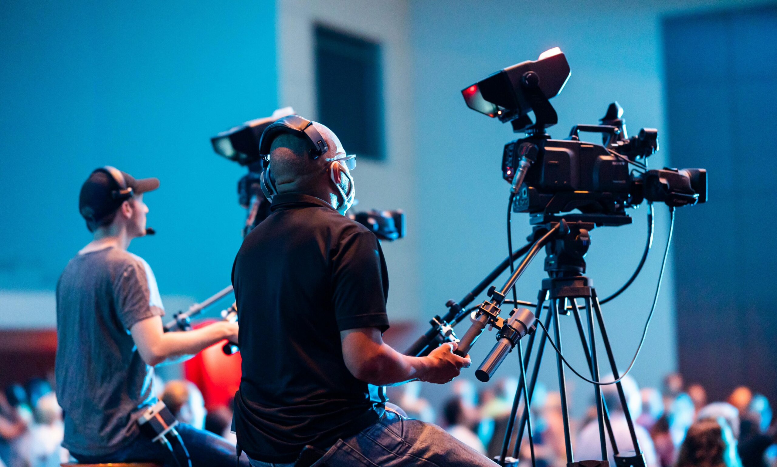 Two cameramen operate professional video cameras at an indoor event with a lively audience.