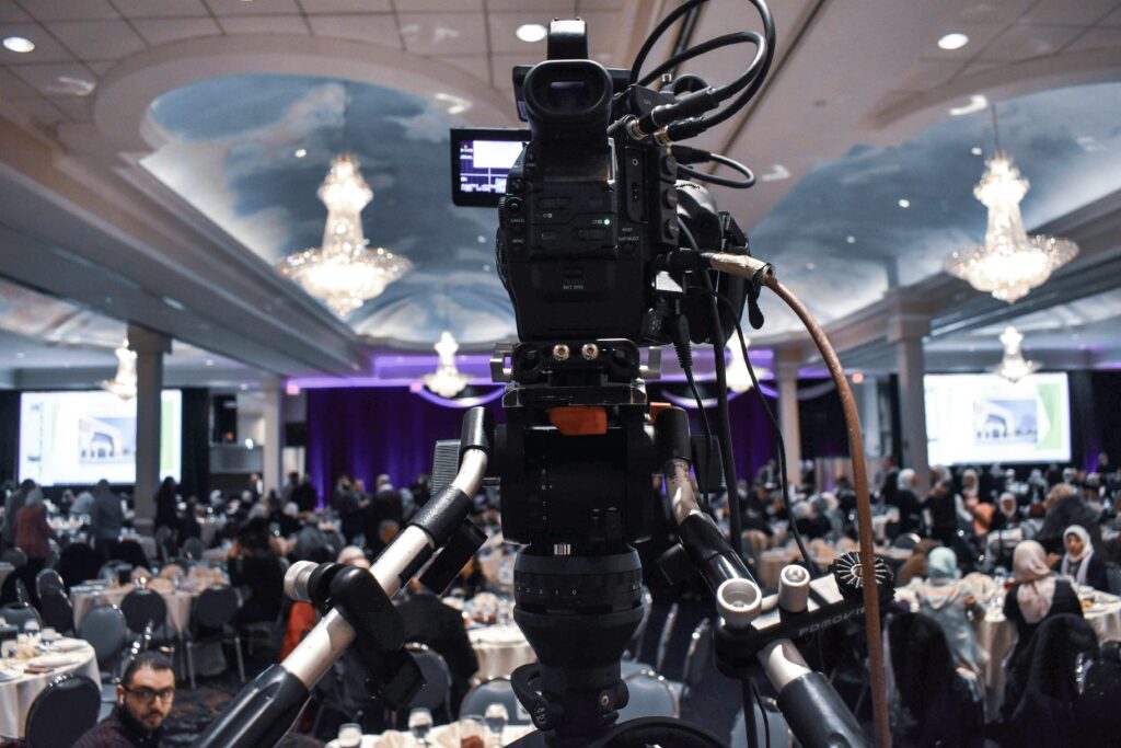Video camera on tripod capturing a large indoor event with chandeliers and busy attendees.