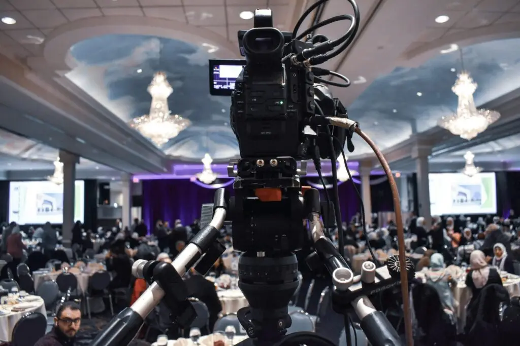 Video camera on tripod capturing a large indoor event with chandeliers and busy attendees.